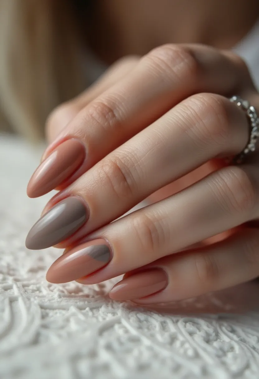 The nail design features a sophisticated and muted color palette primarily consisting of earthy tones, including shades of taupe and beige. The nails are shaped in an almond style, providing a sleek and elegant appearance. The design exhibits a smooth finish which suggests that either gel or acrylic treatments have been used. Two colors are artistically combined on each nail, creating a subtle yet eye-catching diagonal split design pattern. The overall aesthetic is understated, making it suitable for both everyday wear and special occasions, with a particular nod to fall or autumn themes due to the warm and neutral colors.