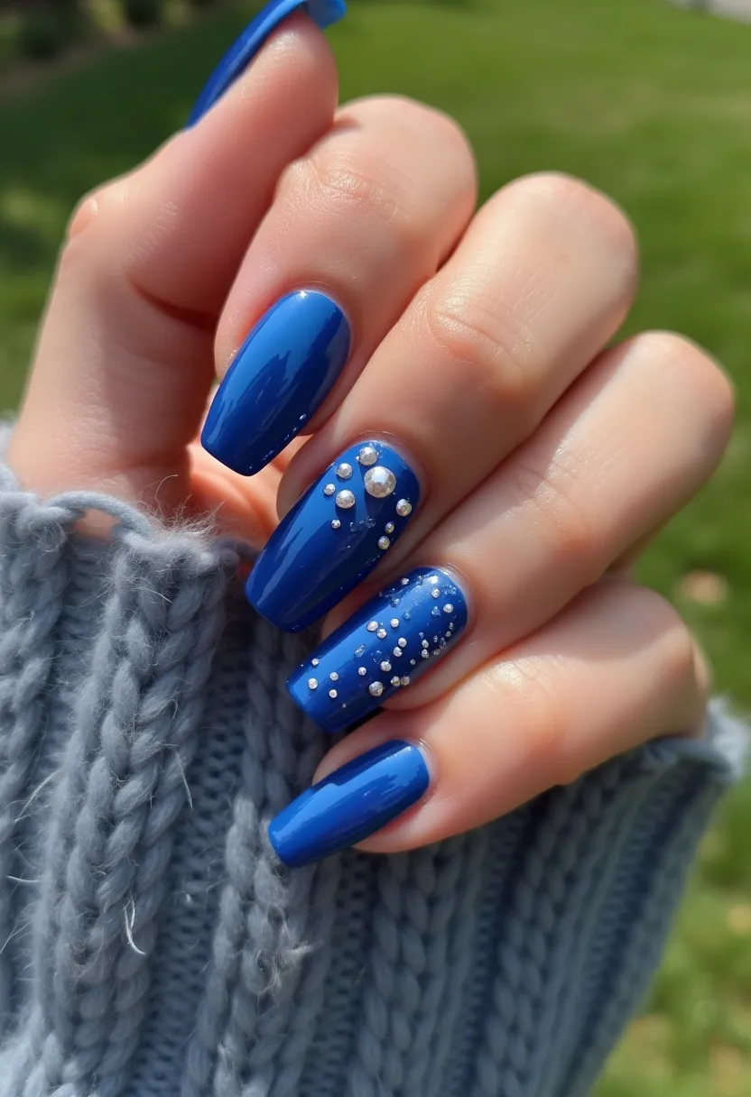 The nail design features a vibrant blue color palette, with nails shaped in a long and rounded square form. The chosen treatment appears to be a glossy gel polish, giving the nails a shiny finish. Two of the nails are adorned with intricate decorations: small rhinestones and pearls are arranged in a scattered yet deliberate pattern. These embellishments add a touch of elegance and sophistication to the otherwise simple design. The look is contemporary and can suit various occasions, boasting both a playful and chic appeal. The sparkle from the decorations might suggest it's suitable for evening events or special celebrations, though it retains a stylish simplicity for everyday wear.