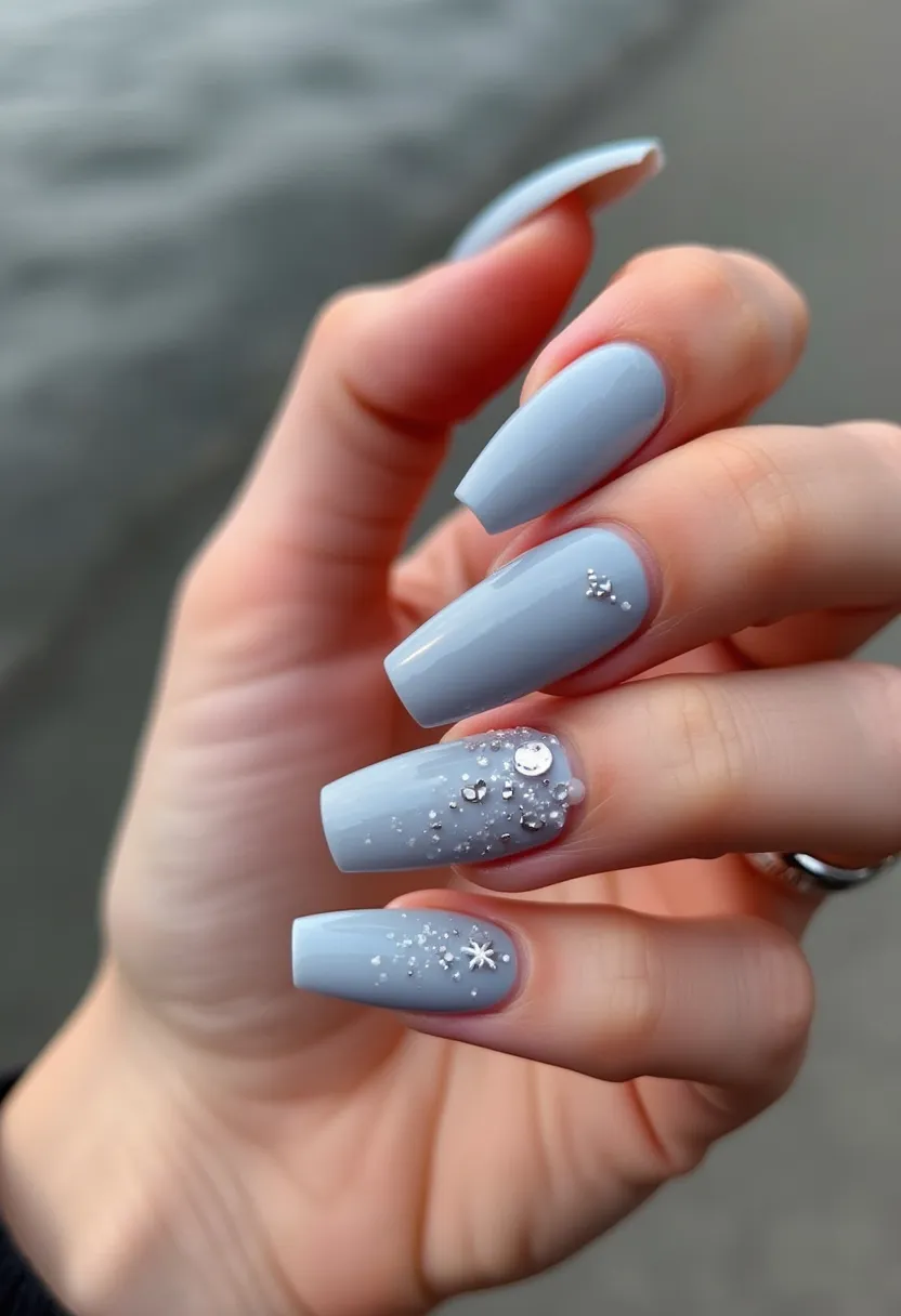 The nail design showcases a soft, pastel blue color palette, creating a serene and elegant look. The nails are long and coffin-shaped, providing a stylish and modern appearance. An intricate pattern adorns two of the nails, featuring small silver and white embellishments, including delicate snowflakes and dots, giving the impression of a winter theme or holiday season. Some of the decorations appear to be tiny metallic studs and glitters, adding a touch of sparkle and glam to the overall design. This nail art appears to be done with gel polish, as indicated by its glossy finish and durability, common in gel nail treatments. The detailed accents and the color choice make it an ideal design for special occasions during the winter season.