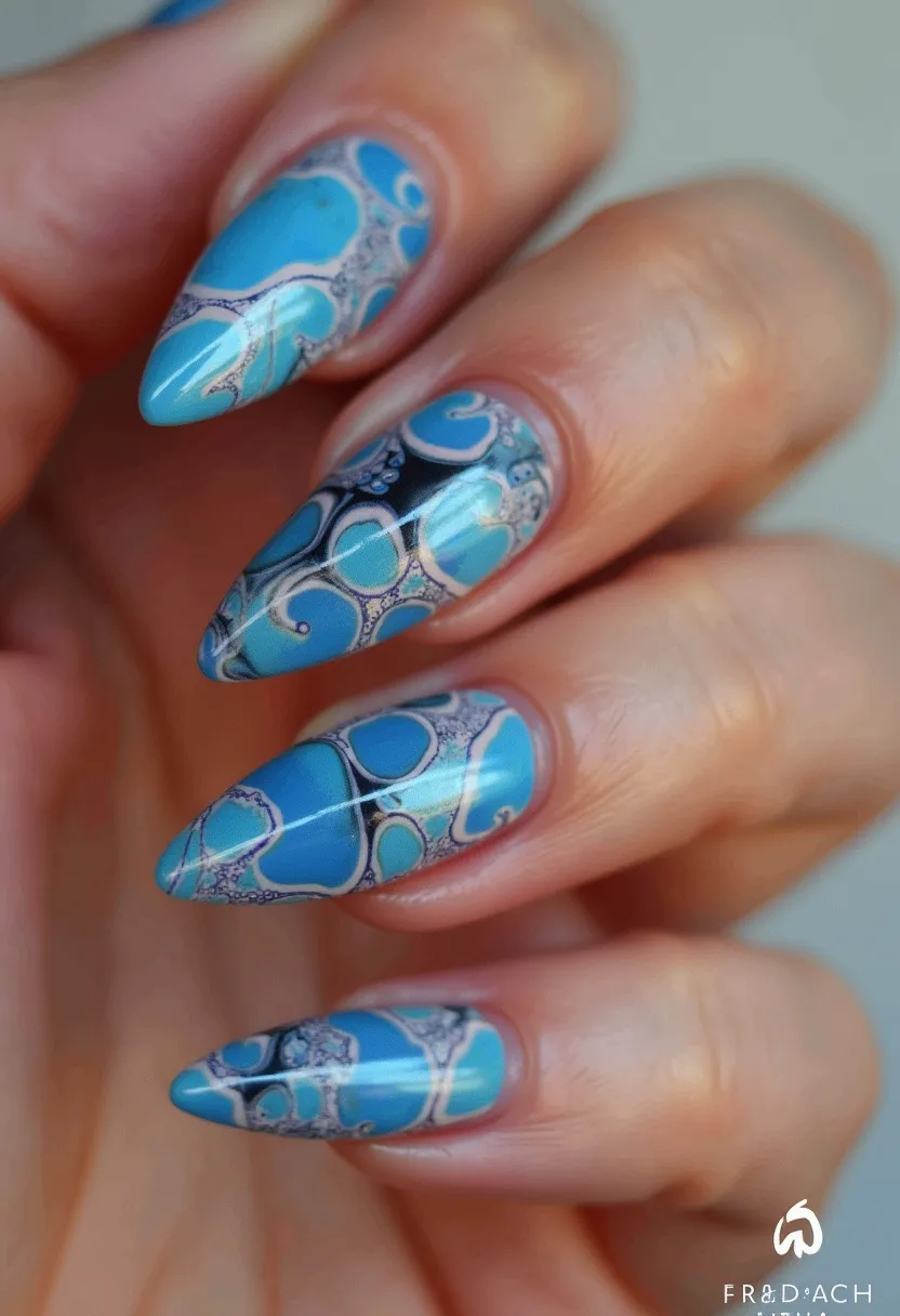 The nail design features a vibrant color palette primarily dominated by various shades of blue, ranging from light sky blue to dark navy. The nails are sculpted into a stiletto shape, offering a sharp and elongated appearance. Intricate patterns adorn each nail, showcasing a marbled or swirling design that interlaces white and grey lines, adding depth and complexity to the blue base. The details suggest the nails are likely treated with gel polish, providing a glossy and smooth finish that enhances the vividness of the colors. This design carries a subtle yet sophisticated elegance, making it suitable for both everyday wear and special occasions, possibly evoking a cool, aquatic theme perfect for summer.