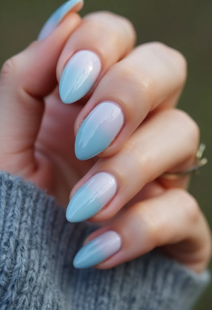 The nail design features an elegant almond shape, painted with a soft and calming pastel color palette of light blue and white. The nails display a gradient or ombre effect where the colors transition smoothly from a milky white at the base to a serene blue at the tips. The high-gloss finish suggests a gel treatment, providing a durable and glossy appearance. This design is simple yet sophisticated, making it suitable for a variety of occasions, with a tranquil hue that can seamlessly transition from winter to spring. There are no additional patterns or embellishments, keeping the focus on the seamless color blend and the flawless finish of the nails.