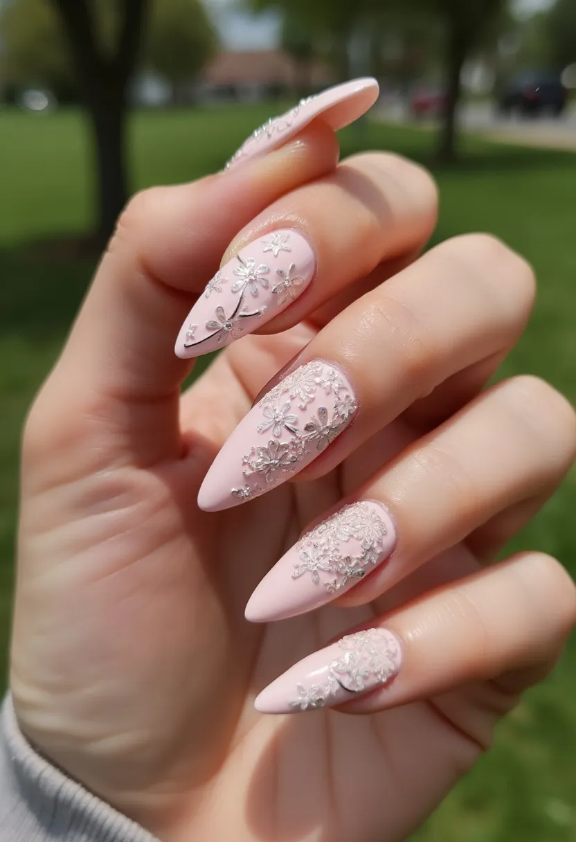 The nail design features a soft, pale pink color palette that provides a delicate and feminine base. The nails are shaped into a sharp point, also known as stiletto style, creating an elegant and dramatic look. Intricate silver floral patterns are delicately placed on each nail, adding a touch of sophistication and charm. The nails are likely treated with gel, given their glossy and smooth finish, which helps preserve the intricate design and extends wear time. The floral patterns and pastel hues suggest a spring or summer theme, making this nail design suitable for special occasions such as weddings or garden parties.