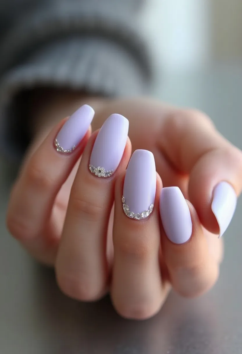 The nail design features a soft lavender color palette, with the nails shaped into a medium-length almond form. The nails appear to be treated with gel polish, providing a smooth and glossy finish. Each nail is adorned with intricate rhinestone decorations placed near the cuticles. The rhinestones vary in size, forming delicate, semi-circular patterns that add an elegant touch to the overall design. This manicure is perfect for events or occasions where a touch of sophistication and subtle glamour is desired.