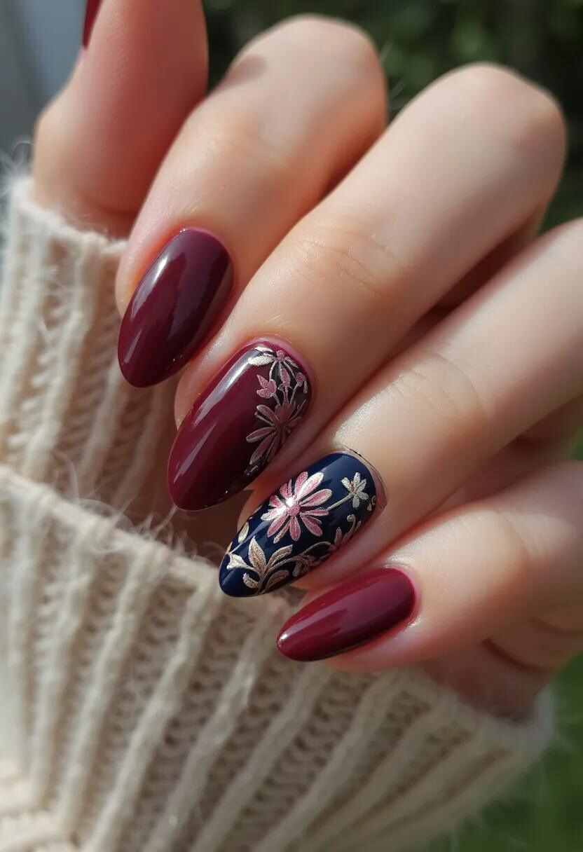 The nail design showcases an elegant and sophisticated aesthetic with a rich burgundy and navy blue color palette. The nails are shaped into a refined almond form, adding to their stylish appearance. Notably, the burgundy nails are accentuated with intricate floral patterns, crafted in a metallic pink and gold hue, adding a touch of glamour and complexity. The floral designs are particularly prominent on the navy blue accent nail, creating a striking contrast that draws attention. This design appears to use gel or acrylic treatment, given its glossy finish and durability. The intricate floral motifs suggest a connection to seasonal themes, potentially evoking a fall or winter ambiance, making them suitable for special occasions like holidays or formal events.