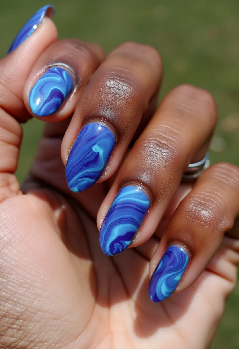 The nail design showcases a marble pattern with shades of blue dominating the palette, ranging from deep royal blue to lighter azure and hints of cyan. The nails are shaped into a medium-length almond or oval form, creating a subtle yet elegant appearance. The intricate marbling effect is characterized by fluid, swirling lines that intertwine to mimic the appearance of natural stone. This detailed pattern suggests a gel manicure, known for its gloss and durability. The design does not explicitly cater to a specific seasonal theme or special occasion but offers a stylish and contemporary look suitable for everyday wear or a sophisticated event.