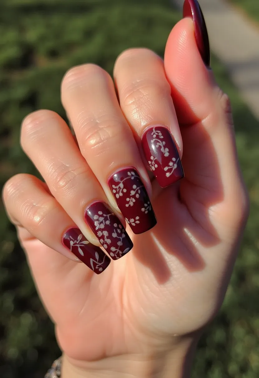 The nail design features a deep burgundy base color, exuding a rich and elegant palette. The nails are medium length with a squoval (square-oval) shape, providing a clean and sophisticated look. Intricate white floral patterns are delicately painted on each nail, adding a feminine and artistic touch. These designs are likely applied using a fine brush or nail stamps, ensuring precision and uniformity. The shiny finish indicates a gel treatment, known for its durability and glossiness. The overall aesthetic suggests a fall or winter theme, perfect for special occasions or festive seasons. The combination of deep color and delicate floral designs creates a balanced and stylish manicure.