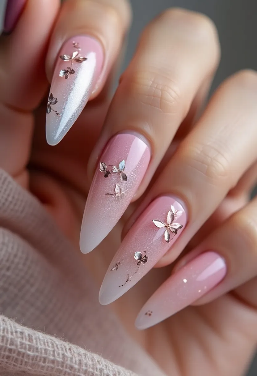 The nail design features a sophisticated ombré color palette that transitions from a soft pink base to a shimmering white tip. The nails are shaped into long, elegant stiletto points, accentuating a refined and striking look. Each nail is adorned with intricate metallic floral decorations, giving the appearance of delicate, three-dimensional silver flowers, which add an element of luxury and elegance to the overall design. The type of nail treatment appears to be a gel manicure, contributing to the glossy and smooth finish observed. This design is perfect for seasonal themes such as spring or special occasions like weddings or formal events due to its elegant and ethereal aesthetics.