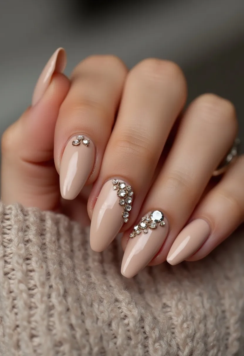 The nail design features a subtle, elegant palette of nude beige polish, providing a sophisticated base color. The nails are almond-shaped, which adds a touch of graceful elongation to the fingers. Intricate decorations include small, strategically placed rhinestones that create a sparkling and luxurious effect, highlighting certain nails more prominently. This design is likely achieved using gel polish, given the glossy and durable finish. The intricate rhinestone detailing suggests care and precision, making the design suitable for special occasions such as weddings or formal events. The overall look combines simplicity with a touch of glamour, perfect for a refined and polished appearance.