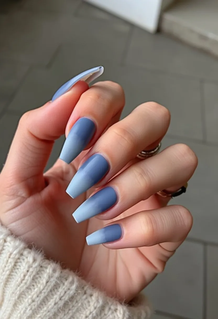 The nail design features long, coffin-shaped nails with a gradient color palette that transitions from a deep navy blue at the base to a lighter, almost sky blue at the tips. The nails have a smooth and glossy finish, suggesting they might be gel-treated. There are no intricate patterns or additional decorations, which helps highlight the elegance of the gradient effect. The overall look is sophisticated and could be ideal for the winter season or any formal occasion due to its cool, subdued color palette and polished appearance.