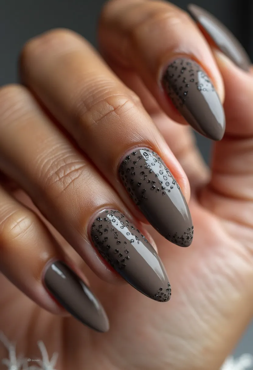 The nail design features an elegant and sophisticated look, predominantly utilizing a taupe or dark taupe color palette. The nails are long and almond-shaped, offering a sleek and elongated appearance. The design includes intricate patterns and decorations where black glitter is delicately sprinkled at the tips and near the cuticles, creating a gradient effect. This design likely incorporates gel polish, as evidenced by the high-gloss finish, which adds a touch of sophistication and durability. The glittery accents provide a subtle yet striking decorative touch, suitable for fall or winter seasons, or even for special occasions such as evening events or festive celebrations.