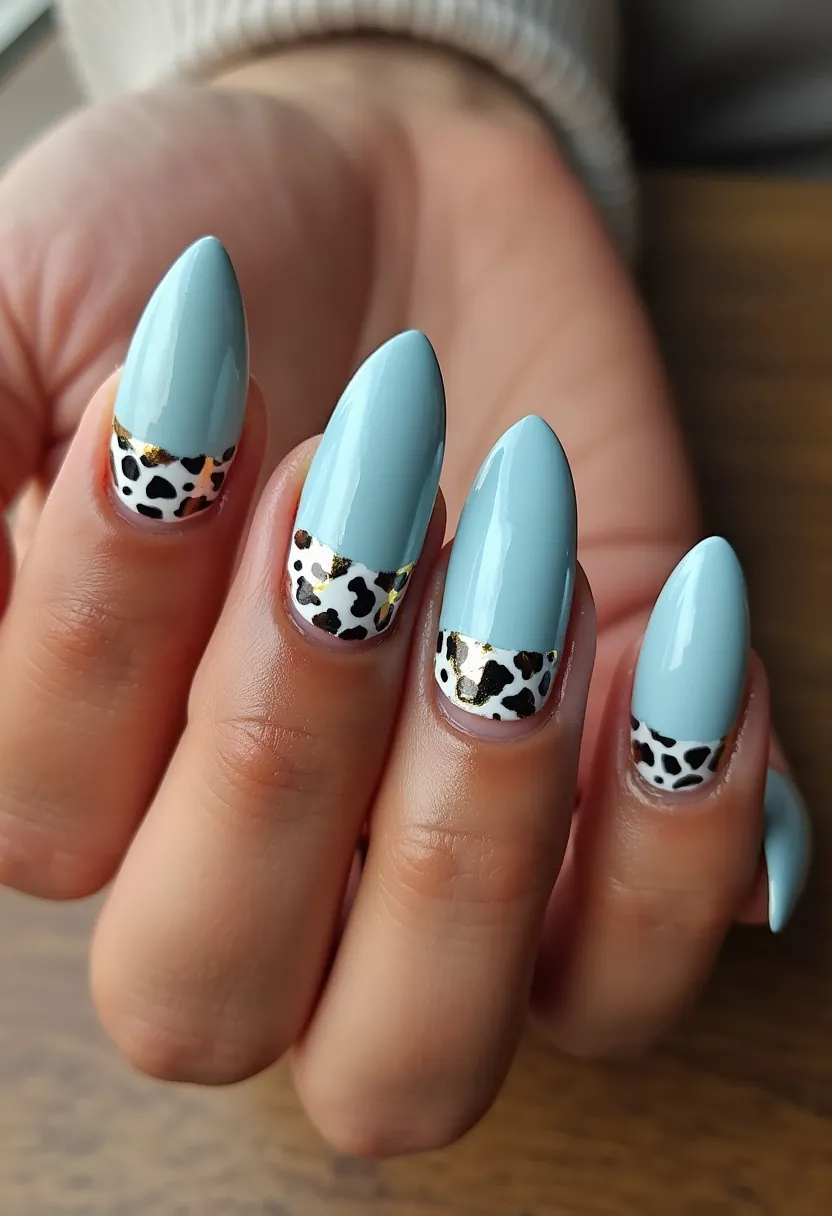 The nail design features an elegant and trendy appearance with a pointed, almond shape. The primary color palette on these nails includes a soft light blue for the main surface area. Intricate patterns include black and white animal print located near the base of the nails, topped with a stripe of metallic gold to add an extra touch of sophistication and glamour. The nails appear to be finished with a glossy coating, suggesting a gel or shellac treatment, providing a smooth and shiny finish. This design integrates a chic and modern aesthetic, making it suitable for various seasons or special occasions where a stylish yet unique look is desired.