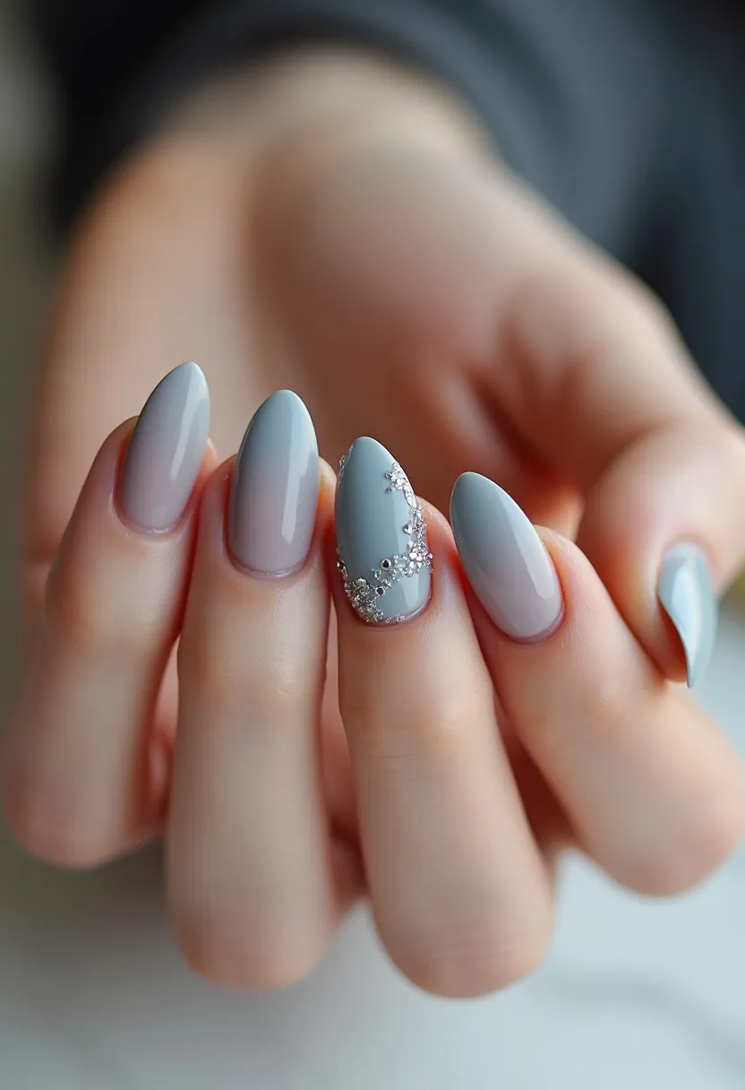 The nail design features an elegant palette characterized by subtle pastel greys and soft blues. The nails are shaped in a chic almond style, which enhances the delicate and feminine appearance. One nail stands out with intricate silver floral patterns and rhinestone accents, adding a touch of glamour and sophistication. The glossy finish suggests a gel or shellac treatment, ensuring durability and a high-shine look. This design could be ideal for a wintertime or formal occasion, given its understated elegance and decorative detailing.