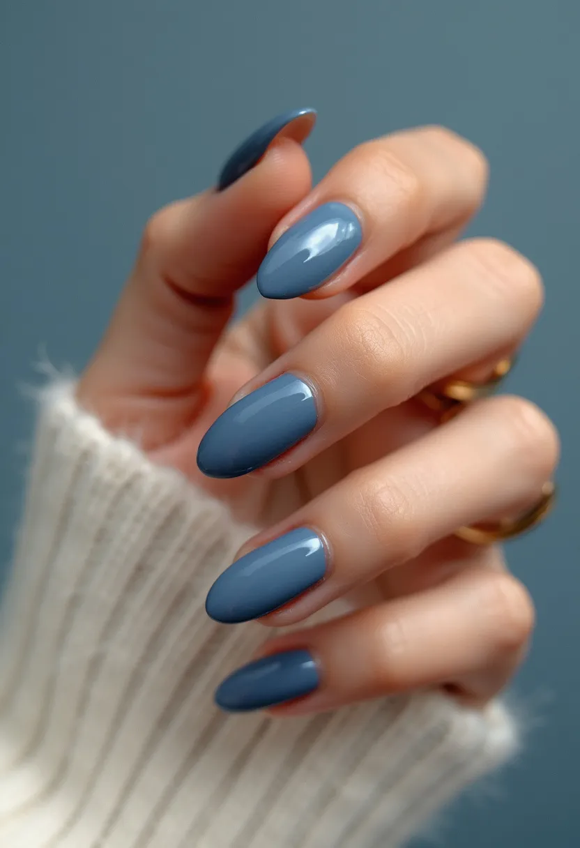 The nail design features a single, solid color palette of a muted, matte blue shade, which evokes a serene and sophisticated look. The nails are shaped into elongated ovals with smoothly rounded tips. The finish is glossy and highly reflective, indicative of either a gel or shellac treatment that provides a sleek and polished appearance. There are no intricate patterns, decorations, or embellishments visible, which keeps the design minimalistic and elegant. This nail style is versatile and suitable for everyday wear, yet classy enough for special occasions, and its cool-toned hue is particularly fitting for cooler seasons like fall or winter.