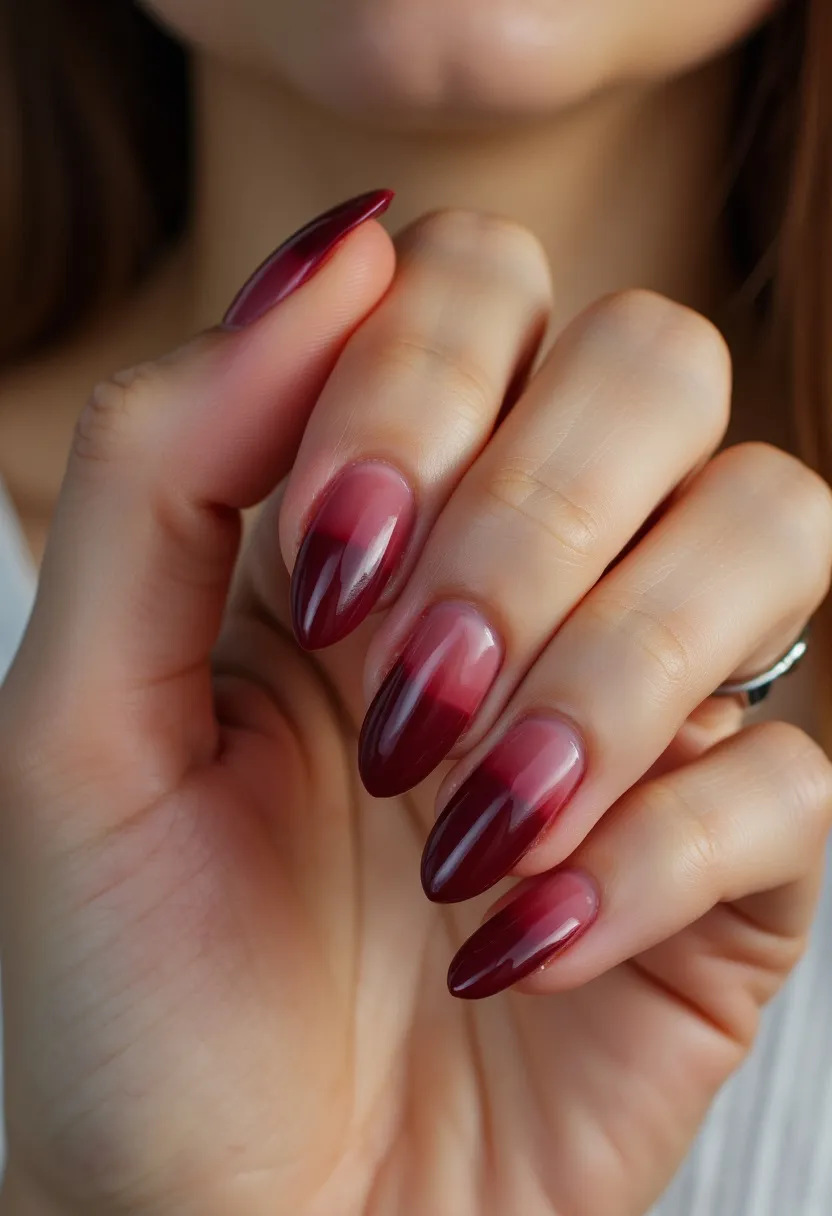 The nail design features a sophisticated color palette primarily consisting of deep reds and maroons, creating an elegant gradient ombré effect that fades towards the cuticles. The nails are shaped into a classic almond style, providing a chic and feminine appearance. This look appears to be achieved with gel polish due to the glossy, smooth finish and durability implied by the design. The translucent gradient offers a visually appealing transition between colors, adding depth and intrigue to the nails. This styling could be suitable for fall or winter seasons, aligning well with holiday festivities or evening events that call for a refined and polished look.