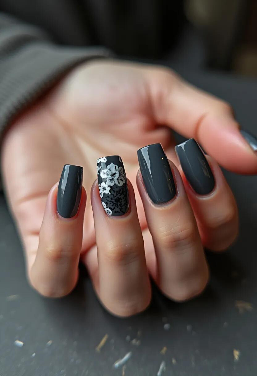 This nail design features a cool gray palette on a squared nail shape, with a glossy finish indicative of a gel treatment. Each nail is elegantly coated in a deep, solid gray color, except for the accent nail which stands out with an intricate white floral pattern. The flowers are delicately detailed, adding a sophisticated and artistic touch to the overall look. This design is versatile and can be seen fitting for both seasonal transitions into autumn or winter, or for a special occasion where a subtle, yet stylish statement is desired. The combination of the neutral color and the floral accent provides a balanced look that is both modern and classic.