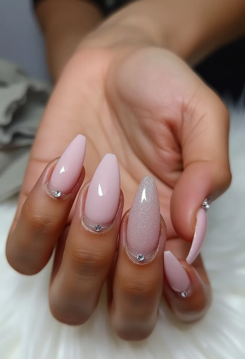 The nail design features an elegant and sophisticated look with a glossy finish. The nails are almond-shaped and have a color palette that includes soft pink shades. One of the nails has a touch of shimmer, adding a bit of sparkle to the overall appearance. Each nail is decorated with a small gem at the base, giving a subtle yet luxurious feel. The likely nail treatment is gel, considering the smooth and shiny surface. This design could be suitable for special occasions or a seasonal theme like spring, due to its delicate and refined style.