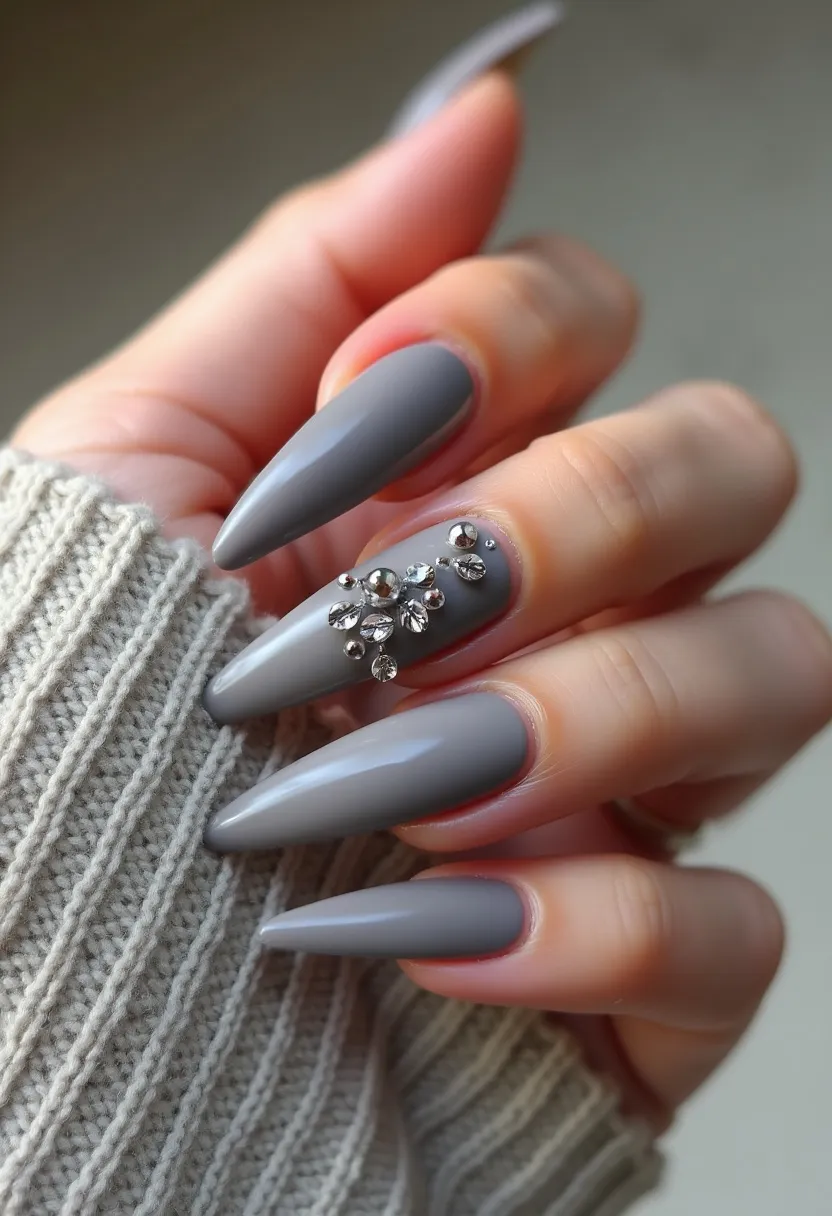 The nail design features a sophisticated palette of cool gray tones, applied uniformly across all nails with what appears to be a glossy finish likely achieved through gel treatment. The nails are shaped in a dramatic stiletto style, characterized by their long and sharply pointed tips. An intricate pattern adorns the ring finger, consisting of a cluster of small, glistening rhinestones arranged in a floral or snowflake-like design, adding a touch of elegance and texture. This design could be suitable for the winter season or upscale events, given its refined color and the sparkling embellishments that evoke a festive or formal occasion.