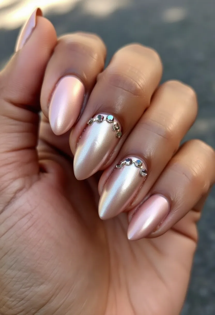 The nail design features a shimmering, metallic light pink or champagne color palette applied evenly across all nails, which suggests a pearlescent finish, likely achieved using either gel or shellac treatment. The nails are shaped into a long almond point, which adds an elegant and sleek look. Each nail is adorned with a delicate line of small, reflective rhinestones near the cuticle area, providing a subtle but glamorous decorative detail. The overall style of the nail design, with its shimmering hue and meticulously placed rhinestones, could suit a festive or formal occasion, such as a wedding or New Year's celebration, adding a touch of sophistication and sparkle.