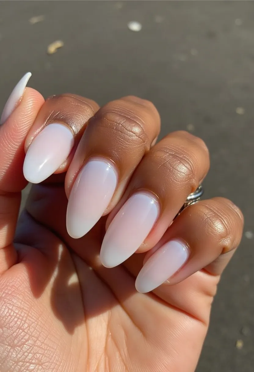 This nail design showcases elegantly shaped almond nails, featuring a subtle and sophisticated gradient of semi-transparent, milky white color which smoothly transitions to a slightly more opaque tip. The nails appear to be treated with a glossy finish likely achieved through a gel manicure, offering a long-lasting and high-shine result. The design is simple yet chic, making it versatile for various occasions, and exuding a clean, modern aesthetic. The overall look suggests a timeless elegance suitable for a variety of settings, from everyday wear to formal events.