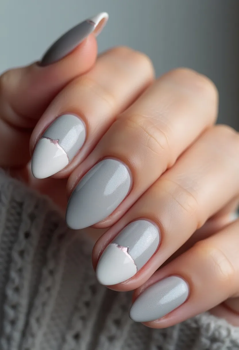 The nail design features an elegant and muted color palette, predominantly using shades of light grey and white. The nails are shaped in a refined almond form, providing a sophisticated look. Two of the nails feature an intricate pattern where the grey and white colors meet in a diagonal split, accentuated with a subtle strip of glittery pink to create a gentle, sparkling division. The finish appears to be a smooth gel treatment, ensuring durability and a glossy effect. The overall design carries a chic and understated elegance, suitable for a winter season theme or any special occasion that calls for a polished yet modest appearance.