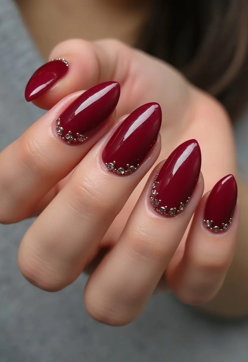 The nail design features a deep burgundy color palette, which gives a rich, elegant look. The nails are shaped into a pointed almond shape, enhancing the overall sophisticated appearance. Each nail is decorated with intricate small rhinestones at the base, adding a touch of glamour and sparkle. The nails appear to be treated with a gel application, as indicated by their glossy and smooth finish. This design could be suitable for special occasions like evening events or holiday parties, given its luxurious and festive look.