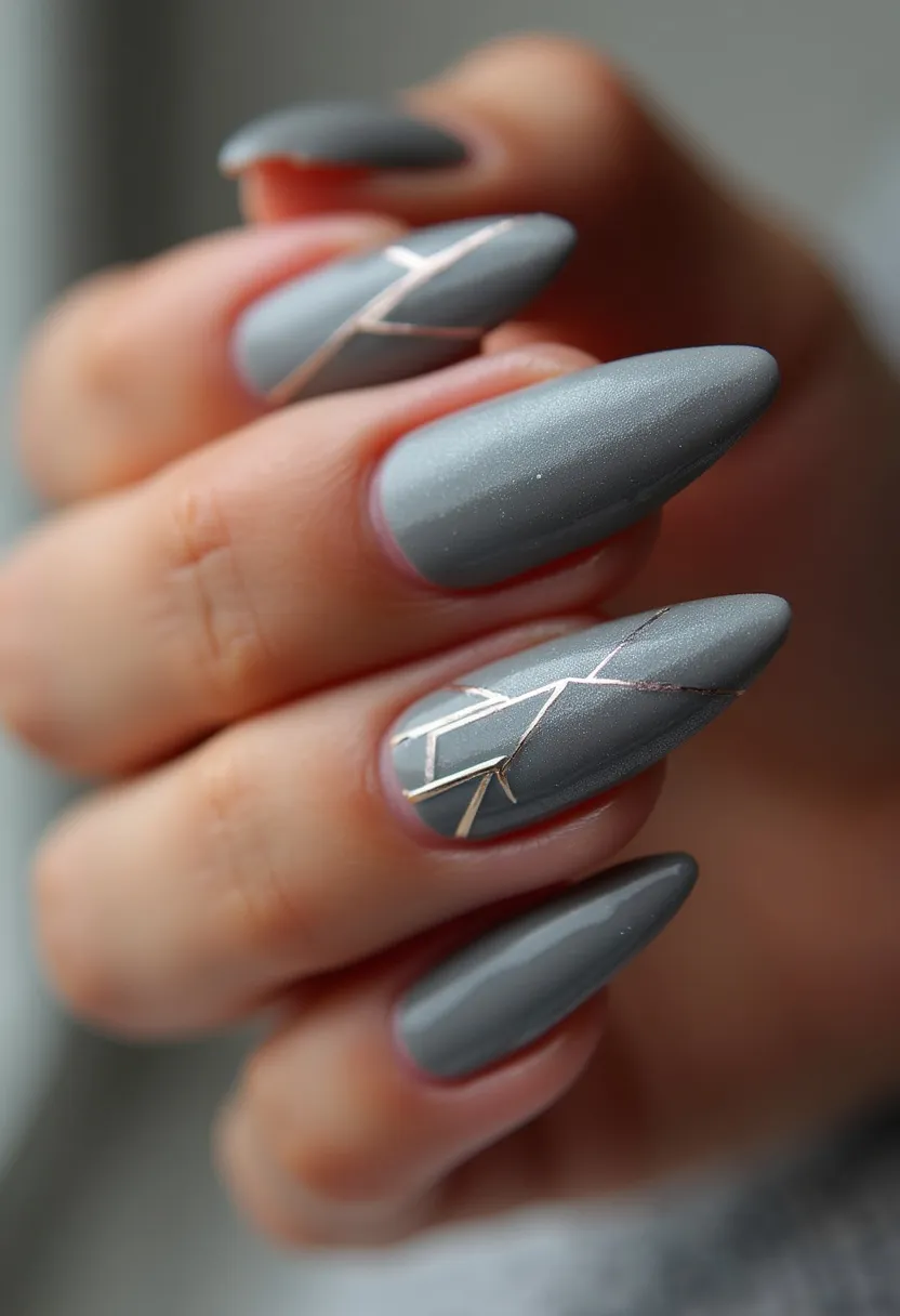 The nails in the image feature a sophisticated grey color palette with a matte finish, indicating a likely gel treatment due to the smooth and seamless application. They are shaped into an almond form, which is elongated and tapered to a softly pointed tip. The nails are adorned with delicate geometric patterns in metallic gold, adding an elegant touch to the overall design. These decor elements include fine lines and shapes positioned at varying angles, creating a modern and stylish look. This nail design could be suitable for various special occasions due to its classy yet understated aesthetic, making it versatile for both casual and formal settings.