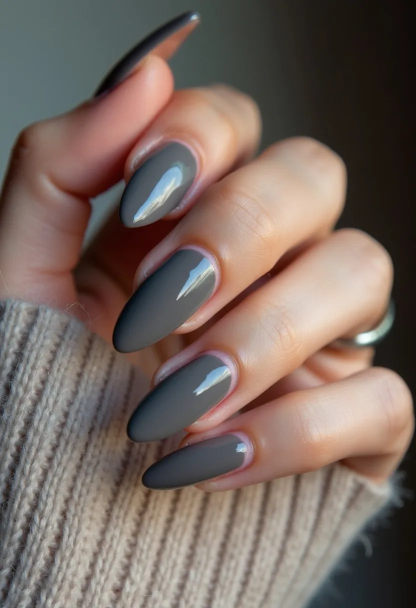 The nail design features sleek, elongated almond-shaped nails coated in a sophisticated grey polish with a high-gloss finish, likely indicative of a gel or shellac treatment. The choice of a single, muted color palette lends the nails a modern and elegant appearance, suitable for various seasons but particularly complementing autumn or winter attire. There are no additional intricate patterns or decorations, relying instead on the simplicity and shine of the solid grey hue to make a subtle but chic statement. The overall look exudes a refined minimalism, perfect for those seeking a polished and professional aesthetic.