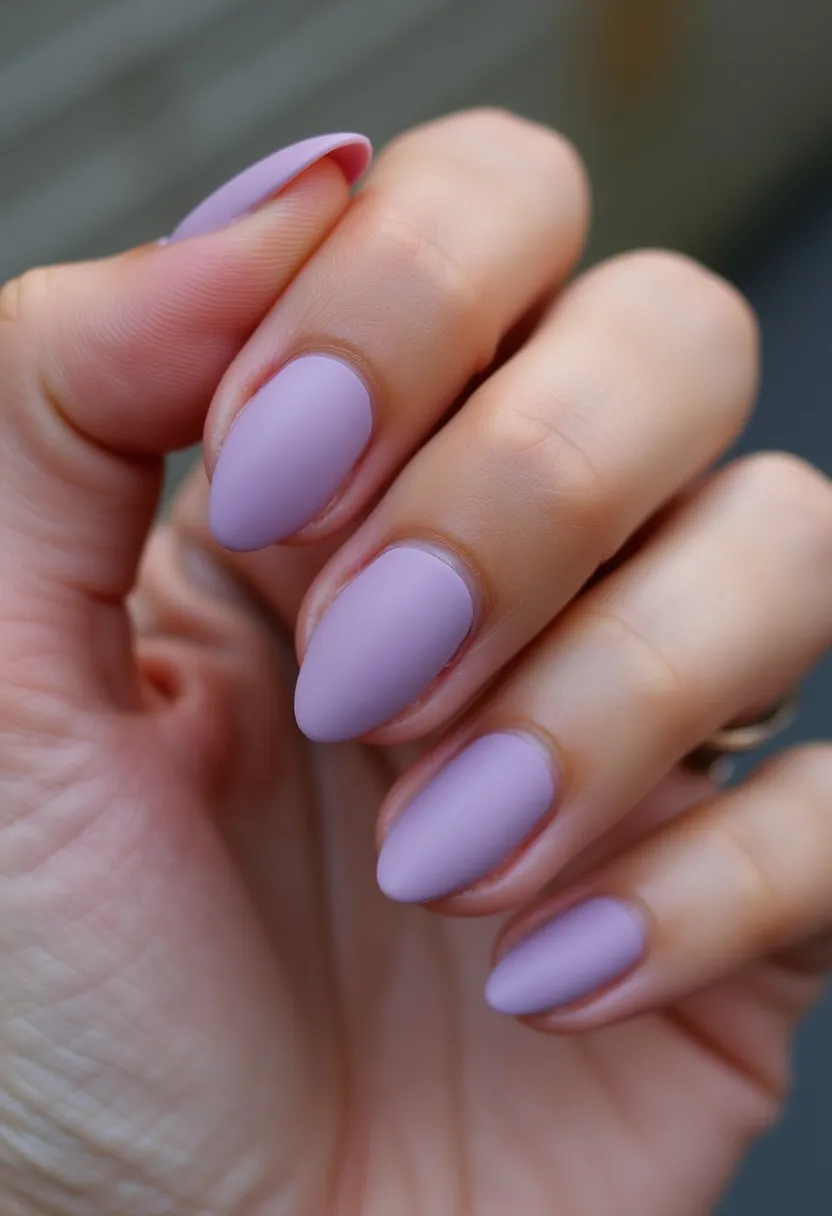 The nail design features a soft lavender color palette, creating a calm and delicate appearance. The nails are shaped in an almond form, offering a refined and elegant look. The matte finish adds a sophisticated touch to the design, without any additional patterns or decorations. This type of nail treatment appears to be a gel manicure, known for its long-lasting and chip-resistant properties. The minimalist and pastel nature of the design makes it suitable for any season, particularly spring or summer, and it can be fitting for various occasions ranging from casual to semi-formal events.