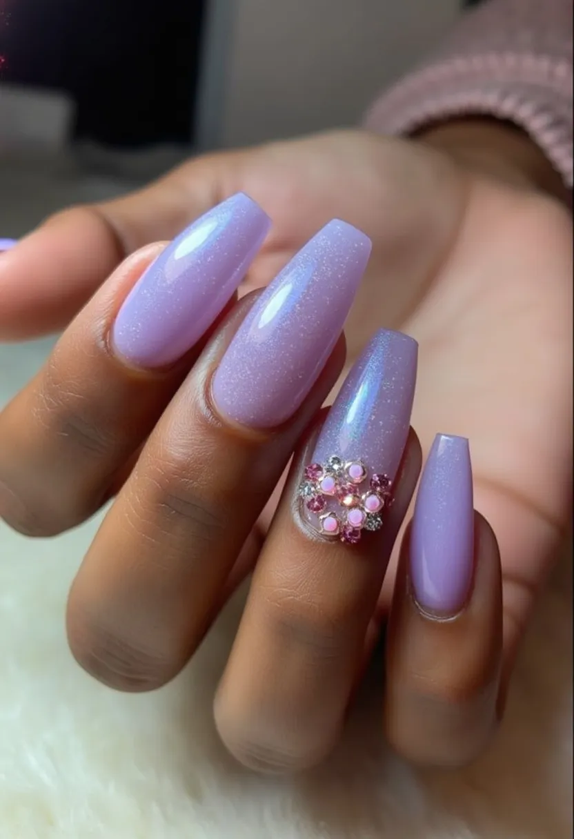 The nail design features a soft, lavender color palette with a glittery sheen, giving the nails a subtle, ethereal appearance. The nails are sculpted into a long, tapered coffin shape, providing a chic and modern look. An intricate decoration is present on the ring finger, showcasing a cluster of embellishments, including small pink and silver rhinestones arranged in a floral pattern. The design appears to use gel treatment, contributing to the smooth, glossy finish. The overall aesthetic is elegant and could be suitable for special occasions or as a stylish, everyday look, with its pastel hue adding a spring or early summer vibe.