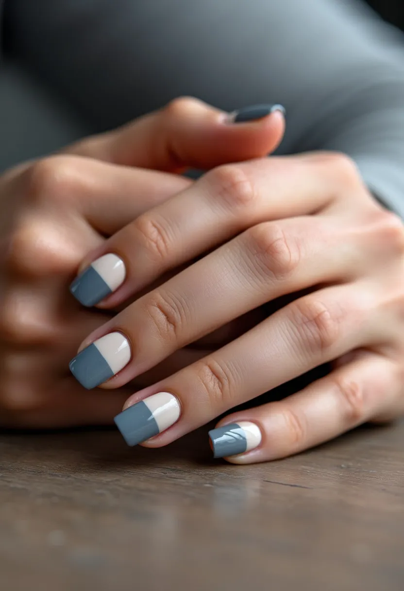 The nail design features a modern and chic aesthetic, characterized by a color palette of muted blue and creamy off-white. The nails are shaped in a natural squared style, allowing for a balanced and sophisticated look. Each nail displays a unique pattern where the creamy off-white serves as a base with a blue block applied diagonally or horizontally across the tips. An additional intricate detail distinguishes the ring finger, which includes a subtle leaf motif etched in white over the blue tip. The finish appears glossy, suggesting a gel treatment for durability and shine. The refined design and cool tones are versatile, fitting for both everyday wear and special occasions, especially suitable for the winter or fall season.