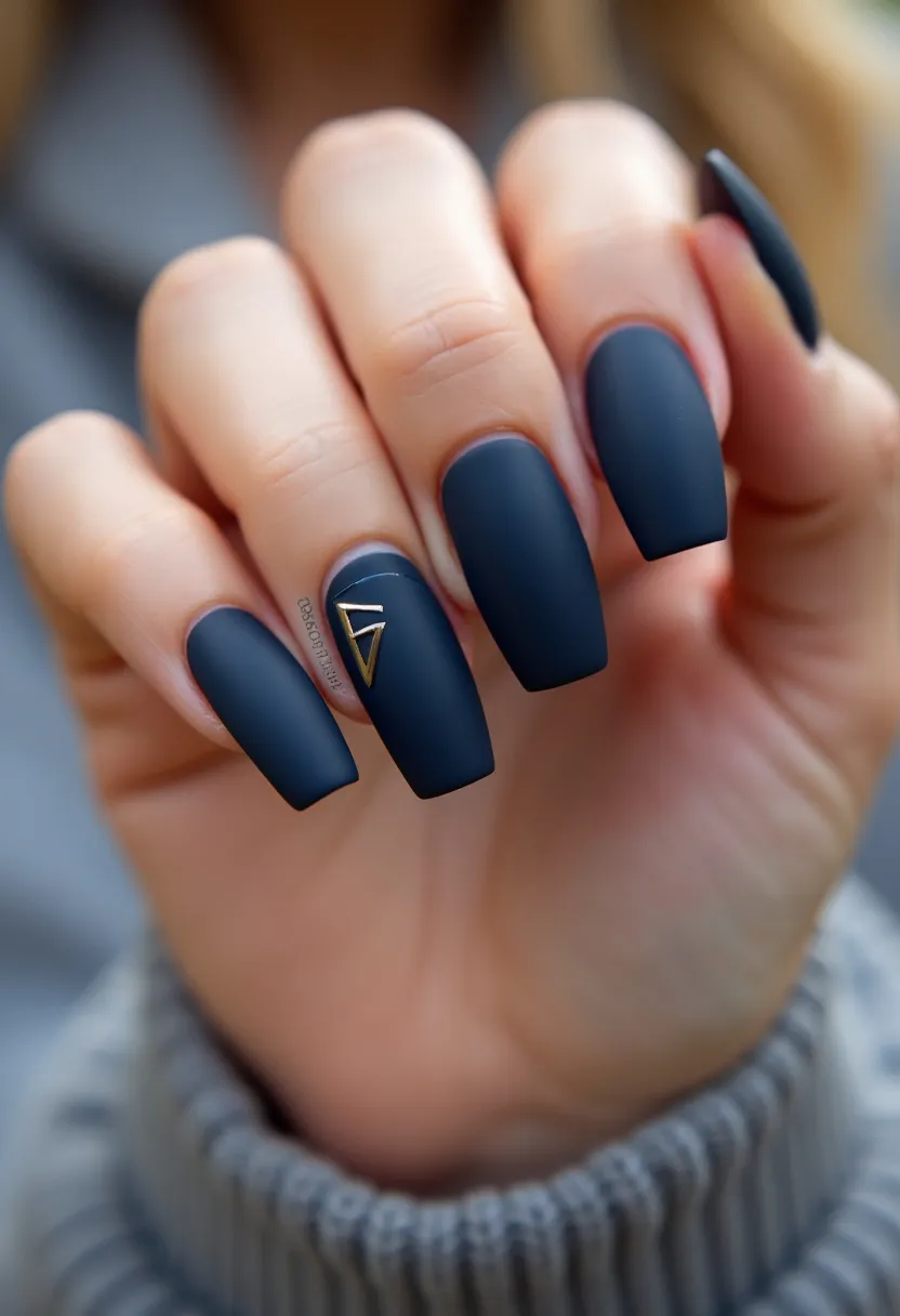 The nail design features a matte navy-blue color palette applied on long, coffin-shaped nails. The nails have a chic and sophisticated look with a smooth finish, suggesting the use of gel polish. An intricate decorative detail is present on the ring finger, consisting of a small gold metallic triangle, adding a touch of elegance and modern appeal. This design is versatile, suitable for both casual and formal occasions, and can be a stylish choice for the fall or winter seasons due to its deep, muted color scheme. The minimalistic yet striking design makes it a standout feature.