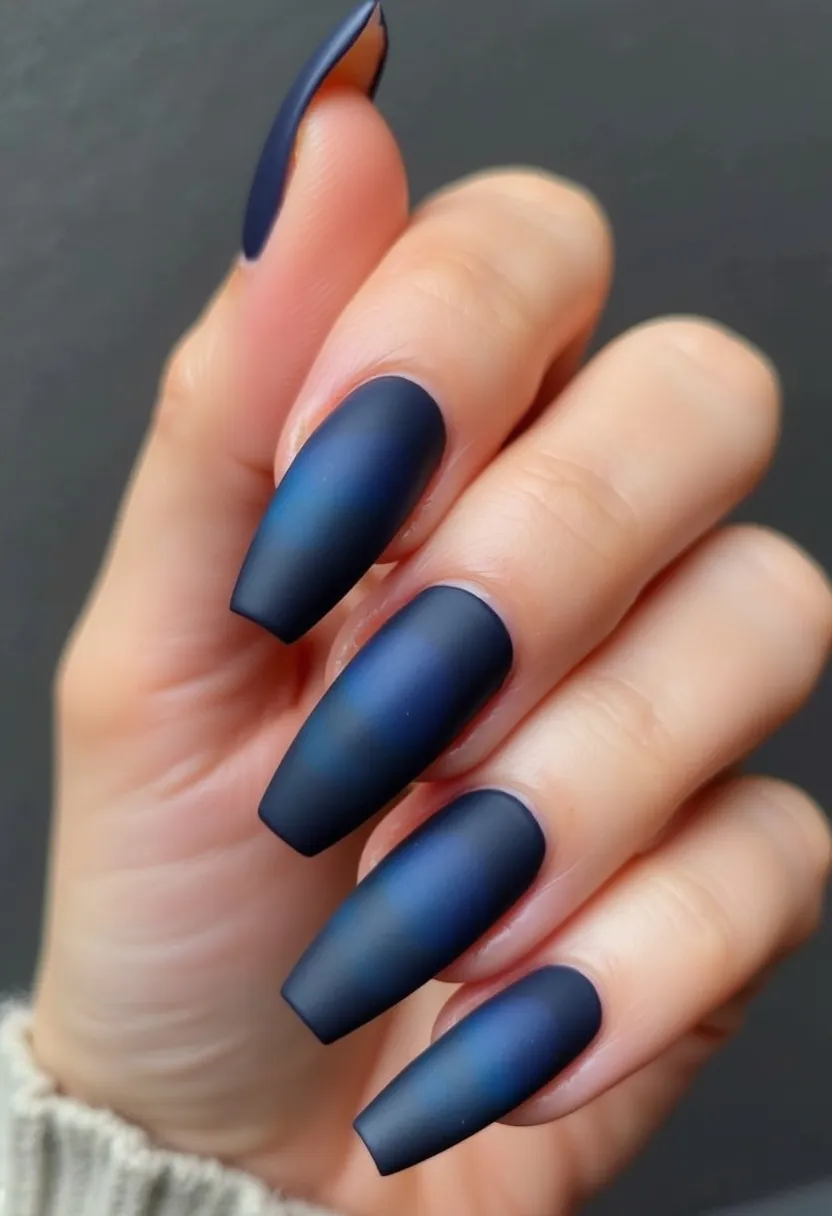 The nail design features a striking matte finish with a deep, midnight blue color palette, showcasing a gradient effect where the color intensifies towards the tips. The nails are shaped in a long, coffin style, creating an elegant and elongating effect on the fingers. The use of gel polish is evident from the smooth, even application and the durable, chip-resistant finish typical of this kind of treatment. This design, devoid of additional patterns or decorations, relies on its rich color and velvety matte texture to make a statement. The choice of a dark, luxurious blue makes this design particularly suitable for the winter season or formal occasions.