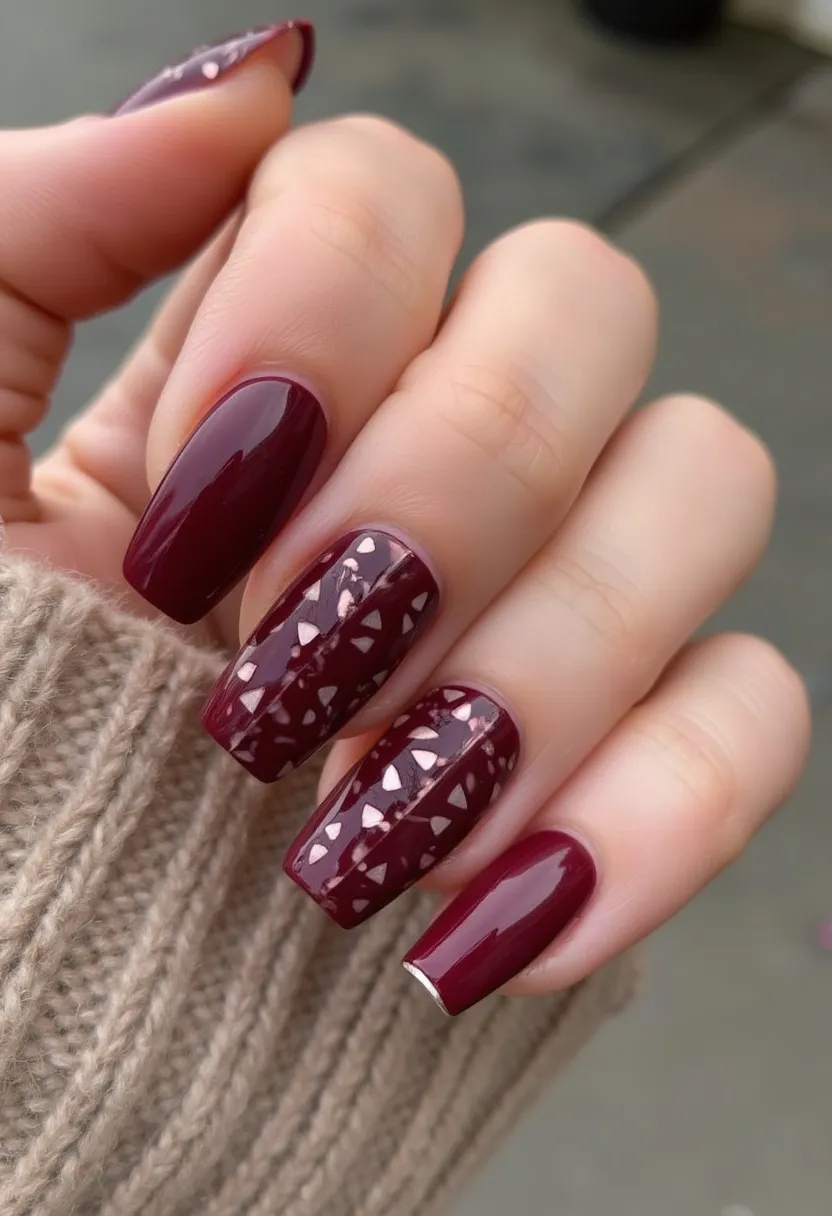 The nail design displays a rich, maroon color palette, exhibiting a sophisticated and polished look. The nails are medium-long and possess a square shape with softly rounded edges. This particular design includes a combination of solid maroon nails and two accent nails per hand featuring intricate triangular patterns. The accent nails are detailed with what appears to be reflective, rose gold geometric shapes embedded within the maroon polish, giving a mosaic-like appearance. The glossy finish suggests the use of a gel or shellac treatment, providing a sleek and durable surface. The overall design exudes a warm, autumnal vibe, suitable for seasonal themes or festive occasions.