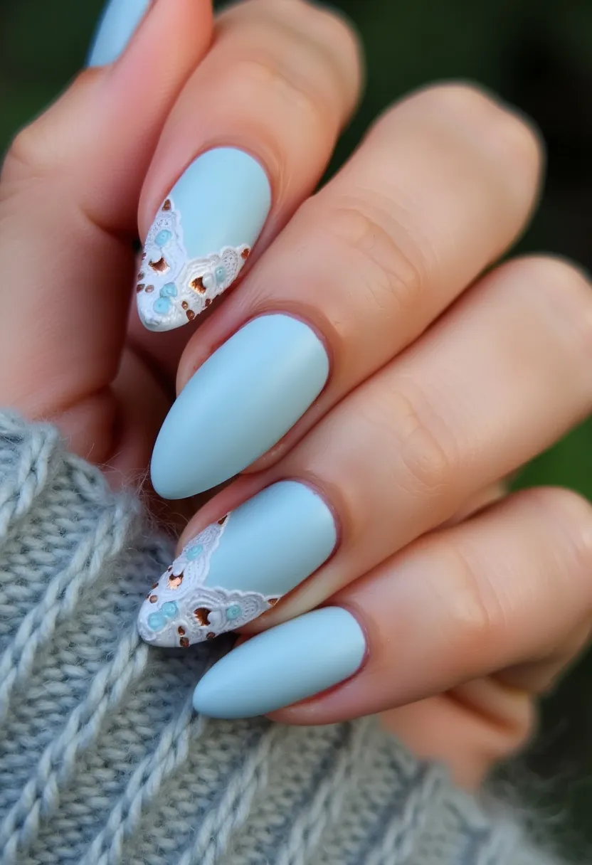 The nail design features a calming light blue color palette, applied as a matte finish. The nails are shaped into a subtle almond shape, providing an elegant and sleek look. Notably, intricate patterns decorate some of the nails, particularly on the ring and index fingers, displaying delicate white lace designs accented with small blue and gold embellishments, adding a touch of sophistication and artistry. The lace-like patterns and hint of metallic details suggest a special occasion or celebratory theme, perhaps suitable for a winter or spring season given the pastel color choice. The nails appear to be treated with gel polish, ensuring durability and a smooth finish.