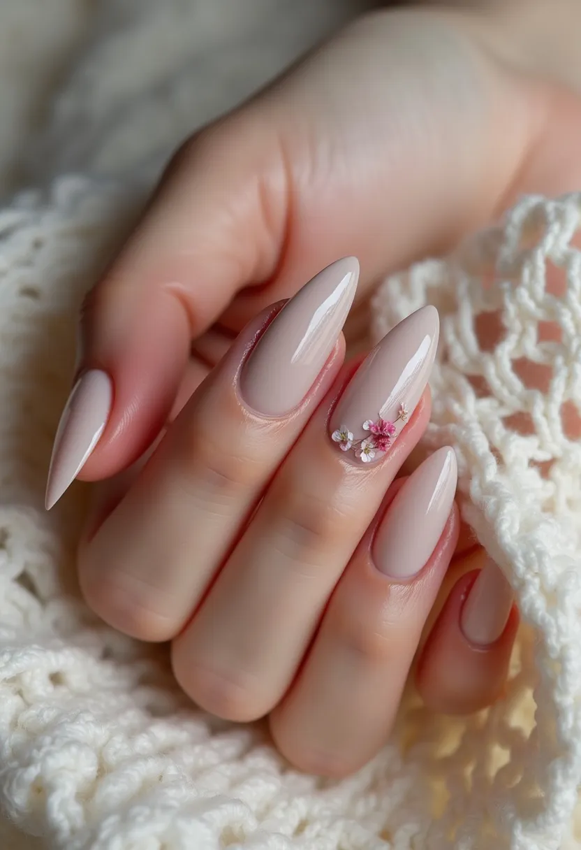 The nail design features a nude color palette with a delicate, cream base that is minimalistic yet sophisticated. The nails are shaped into a sleek almond style, providing a refined appearance. A standout element is the intricate decoration on one nail, which includes tiny pink and white floral appliqués, enhanced by small rhinestones, adding a subtle yet elegant charm to the design. This appears to be a gel treatment, given the glossy finish and durability, making it ideal for long-lasting wear. The floral detailing suggests a springtime or romantic theme, perfect for special occasions such as weddings or celebrations.