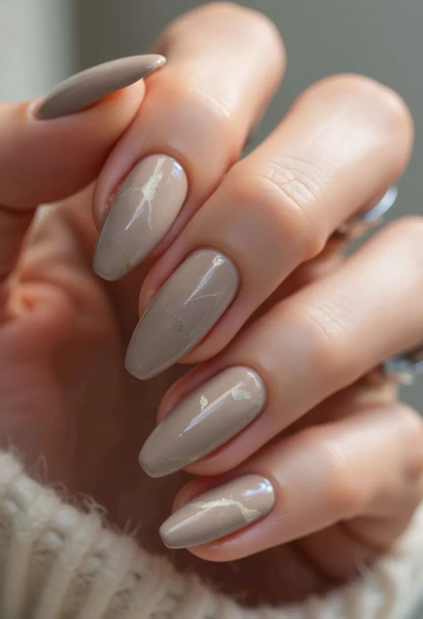 The nail design features a palette of muted, beige-toned taupe with a glossy finish. The nails are sculpted into an almond shape, providing an elegant and elongating effect. The intricate detailing includes delicate, subtle gold leaf accents that are interspersed across the nails, adding a touch of sophistication and luxury to the overall look. This particular manicure appears to utilize a gel treatment, given its high-shine and durable appearance. The design is understated yet chic, making it appropriate for any season or special occasion, particularly formal or festive events.