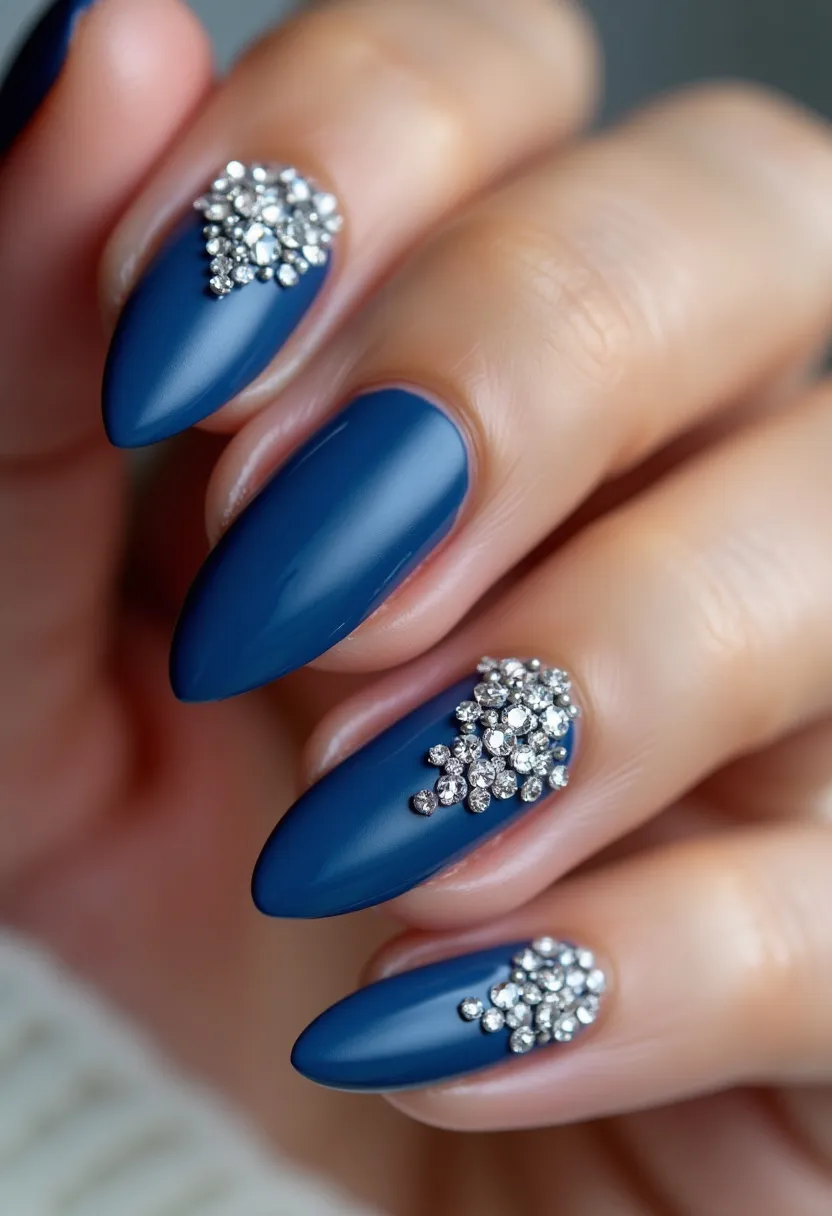 This nail design features a striking color palette of deep navy blue across all nails. The nails are shaped into a long stiletto form, giving a sleek and elegant appearance. Each nail is adorned with intricate patterns of clustered silver rhinestones near the base, adding a touch of glamour and sophistication. The nail treatment appears to be a glossy gel, contributing to the overall polished and refined look. The design elements, including the sparkling rhinestones and rich color, suggest a theme suitable for a formal event or a winter holiday occasion, making the nails a standout accessory for special gatherings.