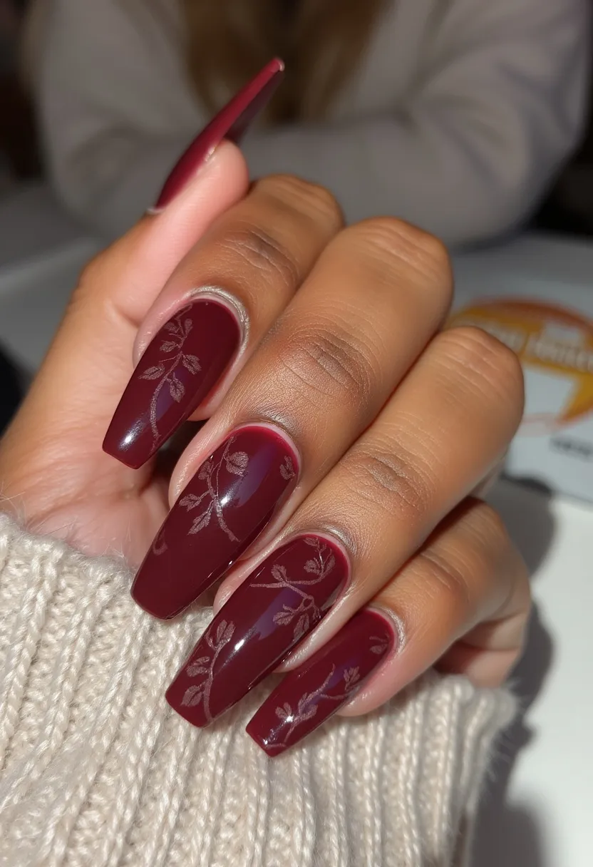 The nail design features a sophisticated burgundy color palette, ideal for fall or winter seasons. The nails are long and have a square coffin shape, exuding an elegant yet bold look. Subtle, intricate leaf patterns in a light, contrasting color are delicately etched onto the nails, enhancing the overall aesthetic with a touch of nature-inspired artistry. The finish appears smooth and glossy, suggesting a gel or possibly shellac treatment, providing durability and shine. This design is both classy and versatile, suitable for special occasions or everyday wear during the cooler months.