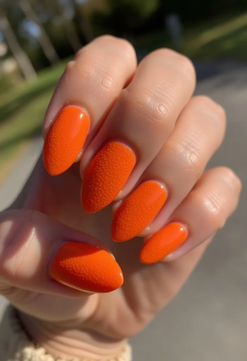 The nail design features a vibrant, solid orange color palette, which is both bold and eye-catching. The nails are shaped into an almond style, with tapered tips that lend a sleek and elegant appearance. This manicure utilizes a blend of glossy and textured finishes, creating a contrast that adds depth and intrigue to the design. The nail treatment appears to be a gel application, given the high gloss and durability indicated by the even, smooth surface. The overall aesthetic suggests a playful and energetic theme, ideal for seasonal celebrations such as Halloween or the fall season when bright and warm colors are in vogue. The unique textured detailing on select nails adds a distinctive touch, setting this manicure apart from more conventional designs.