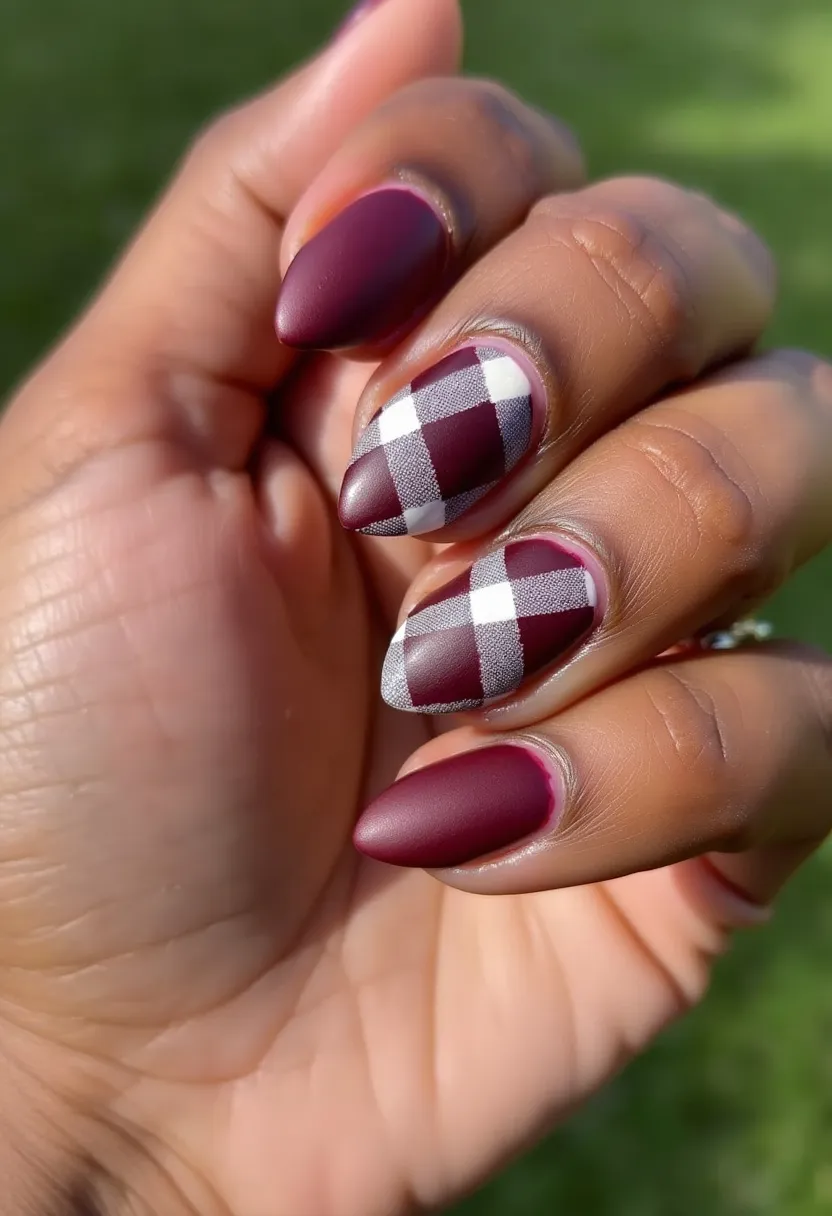 The nail design features a rich, deep burgundy color palette. The nails are shaped in a stiletto or almond style, providing a sleek and elongated look. Two of the nails exhibit an intricate plaid pattern in white and deep burgundy, creating a stylish contrast against the solid burgundy nails. The finish on the nails is matte, indicating either a gel or acrylic treatment with a matte top coat for a smooth, non-glossy appearance. The design has an autumnal or winter theme, making it ideal for the colder seasons or festive occasions. The combination of solid and patterned nails adds a unique and sophisticated flair to the overall look.