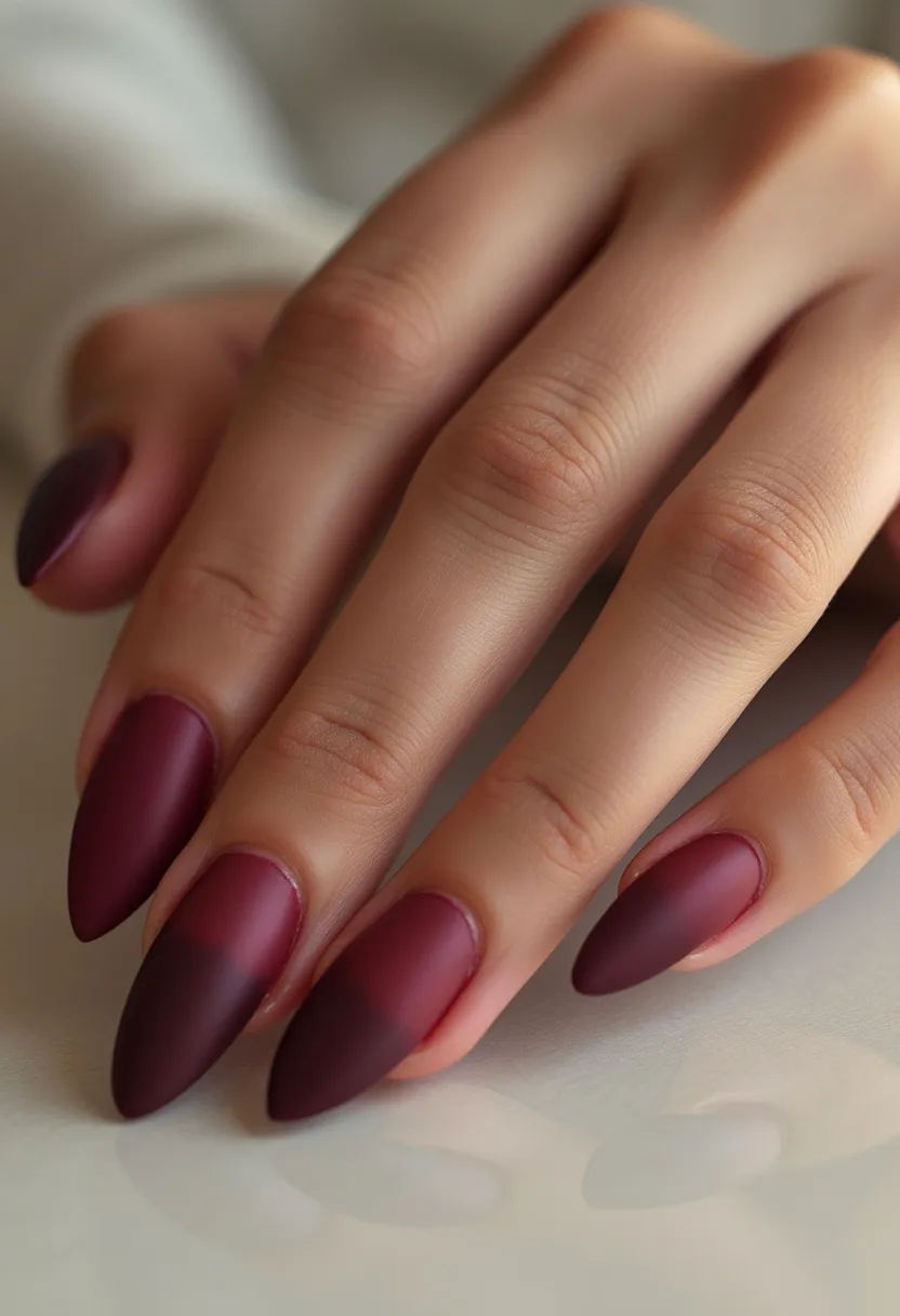 The nail design features a rich, dark burgundy color palette with a matte finish. The nails are shaped into long, sharp stiletto points, creating a dramatic and elegant look. Each nail displays a base color of deep burgundy that appears to slightly shift into a darker gradient toward the tips, providing a subtle ombré effect. The finish suggests the use of gel polish, given its smooth, matte texture that is characteristic of gel nail treatments. The overall design exudes a sophisticated and autumnal or winter vibe, suitable for the colder seasons or special evening events.