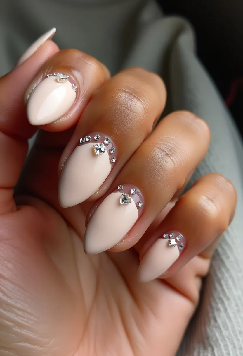 The nails feature an elegant and sophisticated almond shape, coated with a neutral, pale pink or nude polish. The design incorporates intricate embellishments including small rhinestones arranged near the cuticles and a larger, heart-shaped rhinestone in the center of each nail. The rhinestone detailing adds a touch of sparkle and glamour, suggesting a gel or acrylic nail treatment for durability and a glossy finish. This design could be ideal for special occasions such as weddings, anniversaries, or formal events, given its delicate and refined appearance. The overall aesthetic is both chic and understated, making it suitable for a variety of seasonal themes, especially those emphasizing elegance and sophistication.