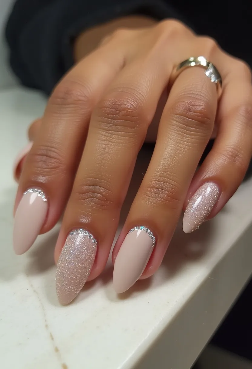 The nail design showcases an almond-shaped style with a sophisticated and elegant color palette consisting of subtle nude tones. The nails appear to be treated with gel for a smooth and glossy finish. Two nails feature intricate decorations: one is entirely covered in glitter, delivering a shimmery, eye-catching effect, while three nails exhibit a minimalist yet glamorous design with a line of rhinestones placed near the cuticles. This manicure exudes a touch of luxury and is suitable for special occasions such as weddings or formal events, or as a sophisticated winter look due to its muted tones and sparkle.