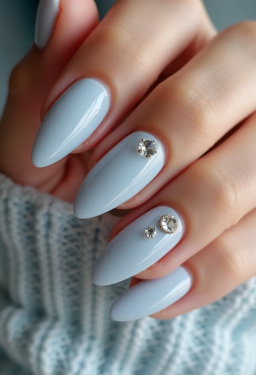 This nail design features a soft, pastel blue color palette, giving a calm and sophisticated look. The nails are shaped in a graceful almond form, enhancing the elegance of the overall design. Adorning the nails are intricate decorations in the form of sparkling rhinestones; one nail exhibits a large, prominent rhinestone, while another showcases a combination of one medium-sized, ornate rhinestone surrounded by a delicate setting, flanked by a smaller rhinestone. These elements add a touch of luxury and glamor, likely achieved through a gel treatment for a smooth, glossy finish. The design exudes a wintry, festive vibe, suitable for special occasions or holiday seasons.