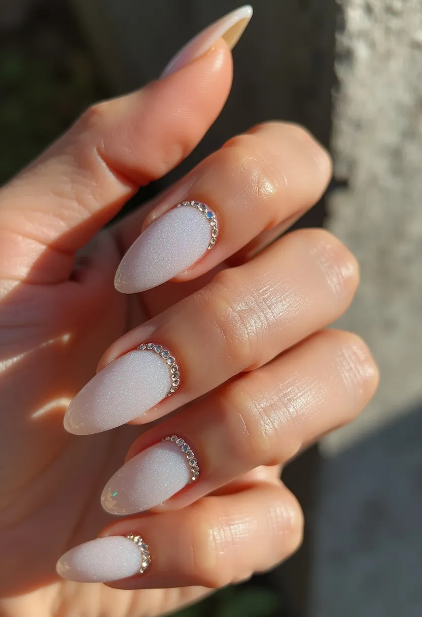 The nail design showcases almond-shaped nails with a subtle, pearlescent white color that gives a shimmering effect. Each nail is decorated near the cuticle with a single row of small, clear rhinestones arranged in a semi-circle, adding a touch of elegance and sparkle. This design is likely achieved using gel polish, as evidenced by the smooth, glossy, and durable finish. The overall look is sophisticated and understated, making it suitable for formal events, weddings, or holiday seasons. The refined use of rhinestones provides a luxurious feel while maintaining a minimalistic aesthetic.