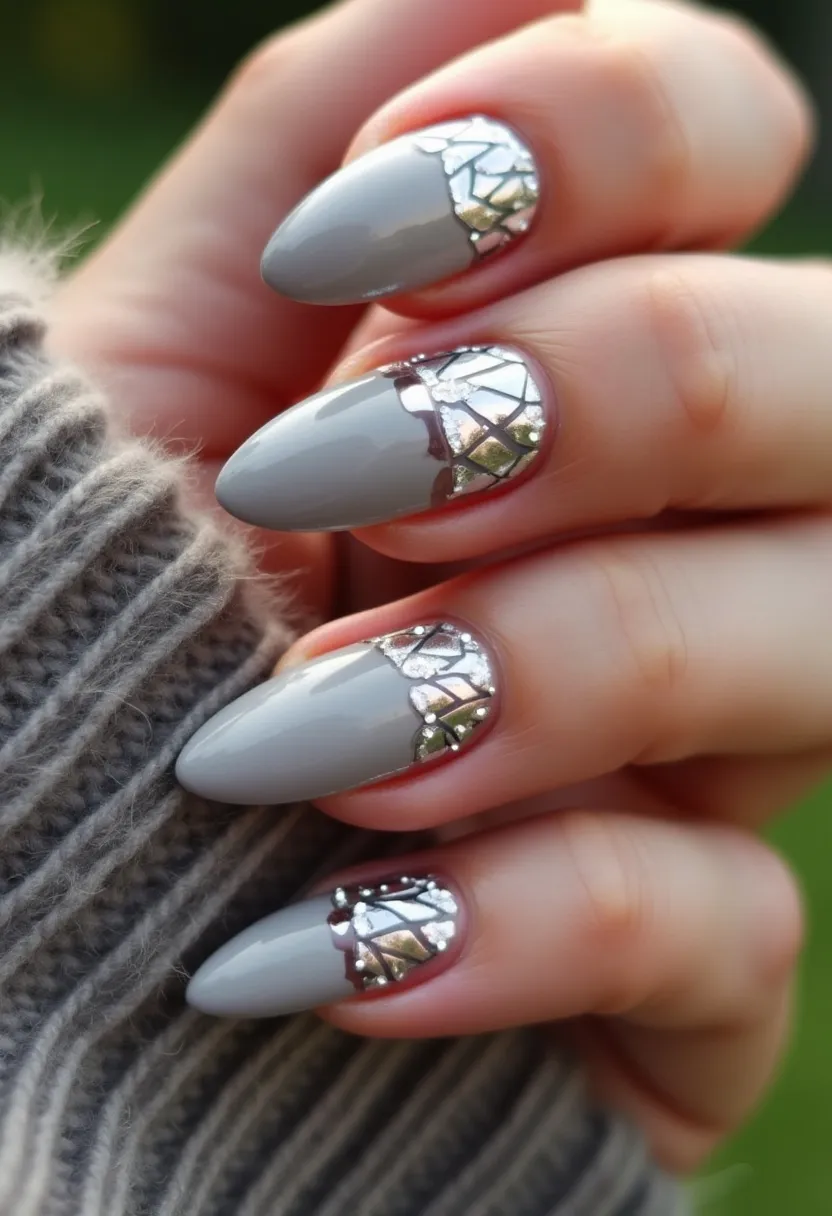 This nail design features a sophisticated and elegant style, perfect for a variety of occasions. The color palette is dominantly a sleek, neutral grey, paired with intricate metallic silver accents. The nails are almond-shaped, which complements the overall modern aesthetic. The design incorporates a sophisticated mosaic pattern near the cuticles, resembling shattered glass or a stained-glass window, adding a unique and chic touch to the nails. The nails likely employ a gel or shellac treatment, providing a glossy and smooth finish that enhances the reflective quality of the metallic accents. This particular design could be ideally suited for a winter season or special events such as holiday parties and formal gatherings, given its stylish and statement-making appearance.