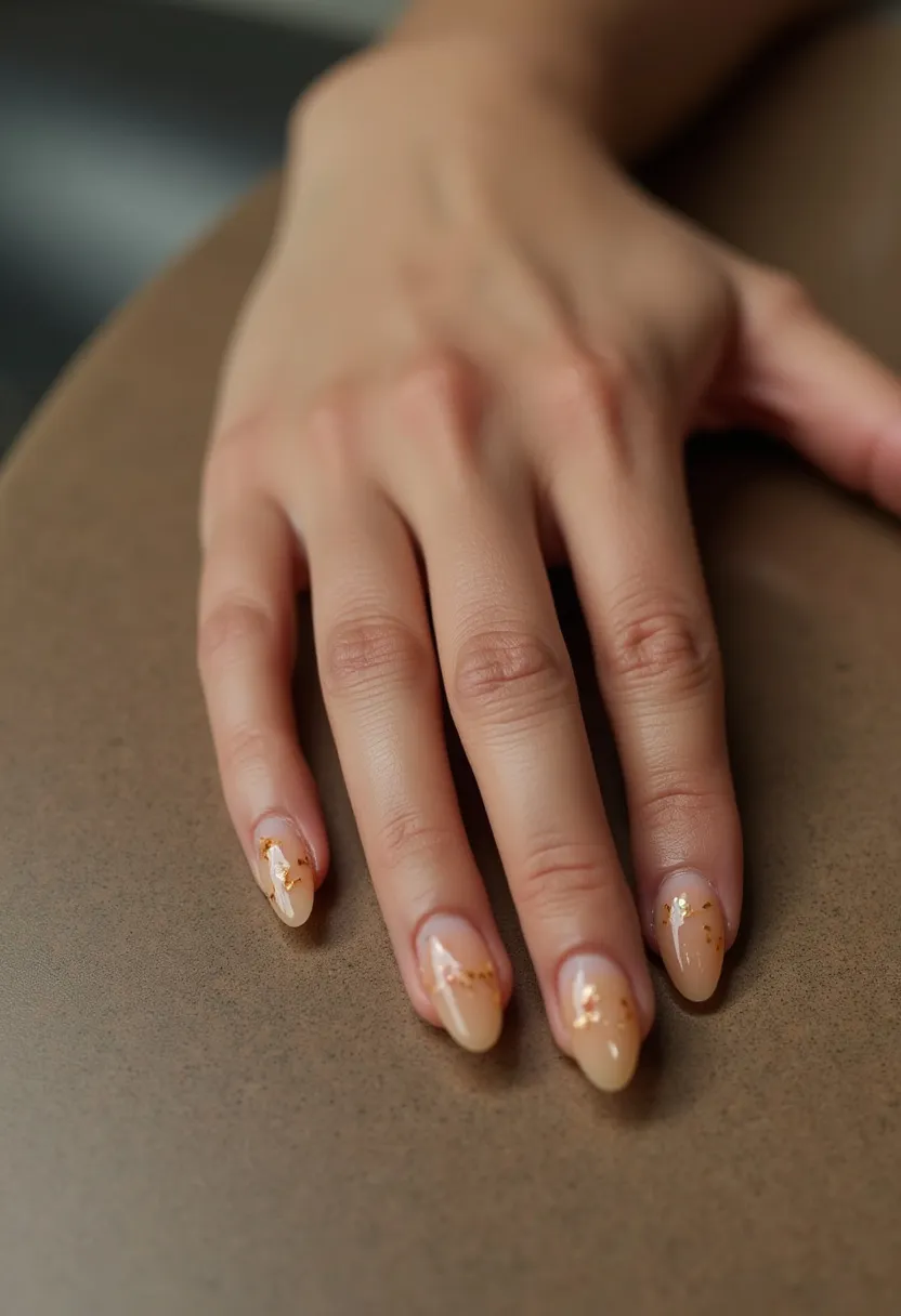 The nail design features an almond-shaped nail with a sophisticated and elegant aesthetic. The color palette includes a warm, nude beige base that seamlessly blends into the natural nail color, creating a sleek and minimalistic look. Intricate gold foil accents are artistically placed on each nail, offering a touch of luxury and glamour without being overly dramatic. The nails likely have a gel or acrylic treatment, ensuring a glossy finish and long-lasting durability. This design is versatile but has a slightly autumnal or festive feel due to the subtle gold detailing, making it suitable for both everyday wear and special occasions.