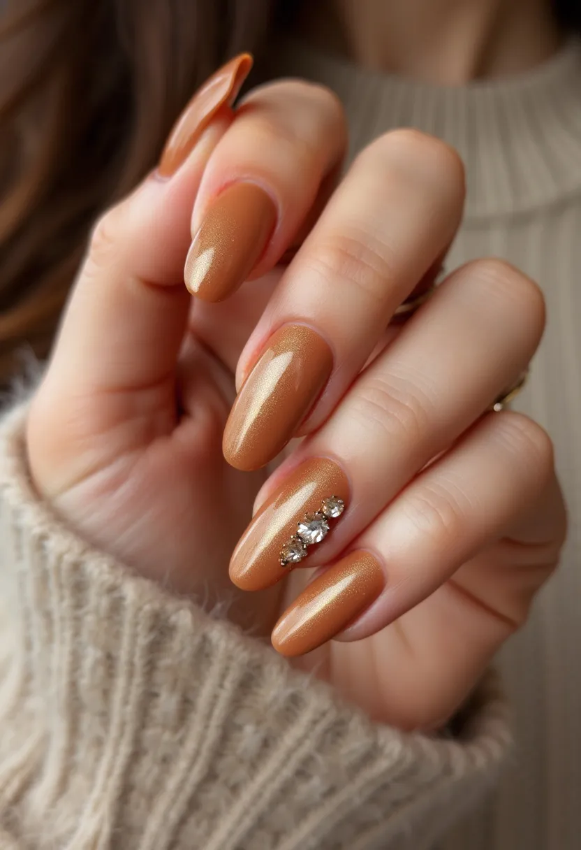 The nail design features a warm, metallic gold color palette that gives an elegant and sophisticated look. The nails are shaped in a medium length, almond style, which is both chic and feminine. The ring finger on each hand is adorned with a line of crystal rhinestones, adding a touch of sparkle and glamour to the overall design. The finish appears to be a gel treatment, providing a glossy and smooth surface that enhances the shimmer of the metallic polish. This nail design is suitable for fall or winter seasons due to its warm tones and would be perfect for special occasions such as holidays or formal events.