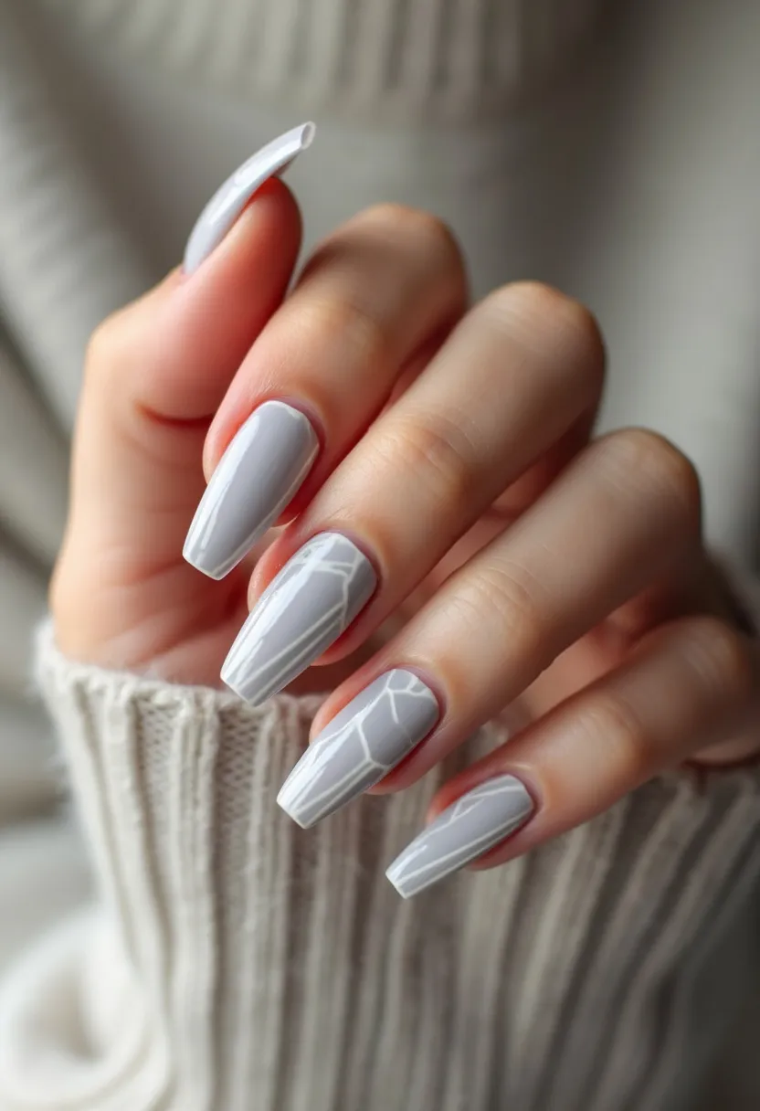 The nail design features a chic and modern look characterized by a muted gray color palette. The nails are in a long, coffin shape which adds to their stylish appearance. The surface of the nails is smooth and glossy, indicative of a possible gel or acrylic nail treatment. There is an intricate white geometric pattern or broken glass effect on some of the nails, adding a unique and eye-catching detail. The design is subtle yet sophisticated, making it suitable for various occasions, from everyday wear to more formal events. The cool gray and white tones suggest a seasonal theme, possibly aligning with winter or a minimalist aesthetic.