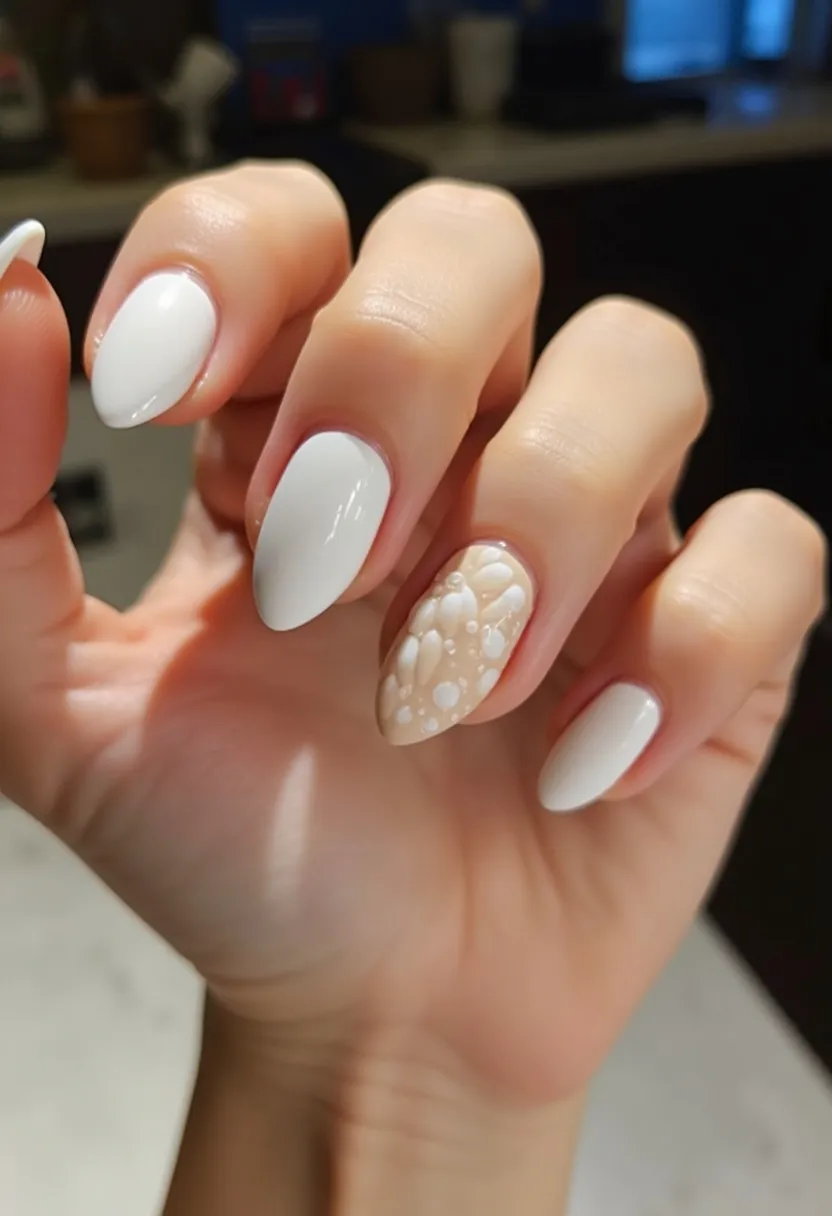 The nail design features a sleek and sophisticated look with a primary color palette of glossy white. The nails are shaped into a soft almond form, which provides an elegant and elongated appearance. The ring finger nail stands out with a distinctive and intricate 3D pattern consisting of white dots and abstract shapes on a creamy beige background, creating a subtle yet effective contrast with the otherwise uniform white nails. The nails seem to be treated with gel polish, given their smooth and shiny finish. This design exudes a classy vibe suitable for bridal occasions or formal events, aligning well with a minimalist yet chic aesthetic.